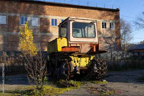 Abandoned tracked vehicle with yellow exterior stands near a brick structure, surrounded by overgrown plants under clear blue sky