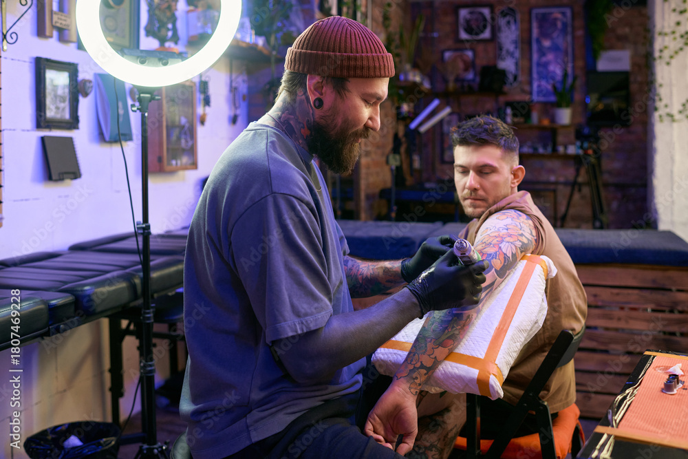Obraz premium Caucasian young adult man tattoo artist working on forearm of Caucasian young adult man client sitting in tattoo studio, both focused on process, tattoo equipment visible in scene