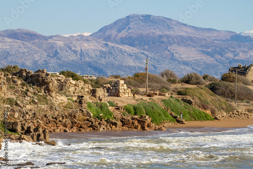 Fototapeta Naklejka Na Ścianę i Meble -  Storm waves crashing on a sandy beach with ancient fortress ruins in Side. A fisherman silhouetted against the dark sea with mountains under blue sky. Mediterranean coast, Turkey.

