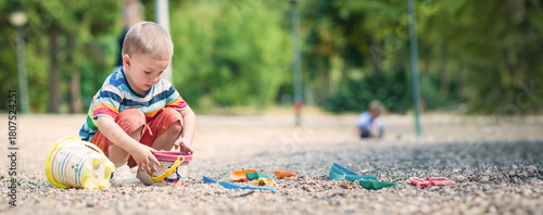 A young child is focused on playing with colorful toys while sitting on soft gravel in a vibrant park. Other children can be seen enjoying the space around them. Place for text
