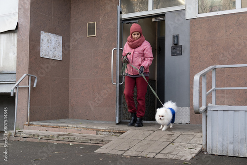 Woman leading white Pomeranian dog on leash out of residential building entrance with a ramp, urban pet care routine, daily responsibility, or veterinary service context