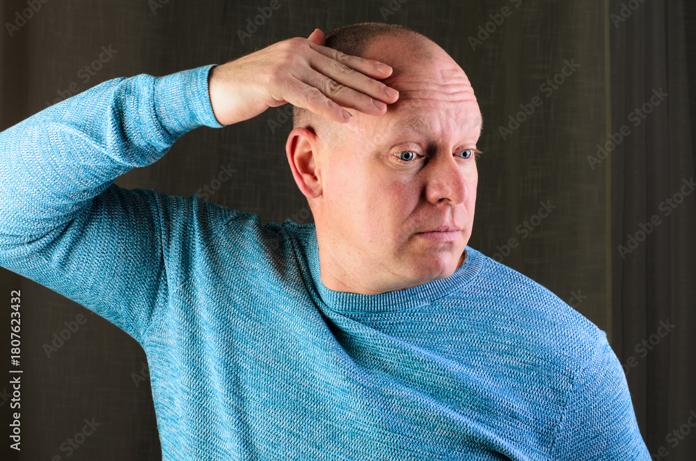 Obraz premium A bald man in a blue sweater is shielding his eyes, perhaps from the sun or deep in thought
