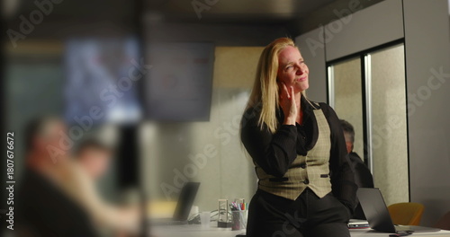 Blonde businesswoman with preoccupied expression gazing into distance in office, reflecting on future while team works in background near conference table