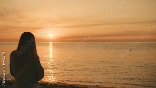 A person stands on the beach, watching the sun set over the calm ocean waters, reflecting warm hues across the sky during a peaceful evening.