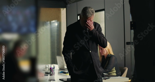 Businessman deeply stressed with hands over face while standing in meeting room as female colleague works in background on laptop during corporate crisis