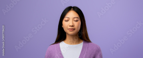 A woman stands with her eyes closed, embracing a moment of mindfulness. She shows a serene expression while wearing a cozy light top and a purple sweater.