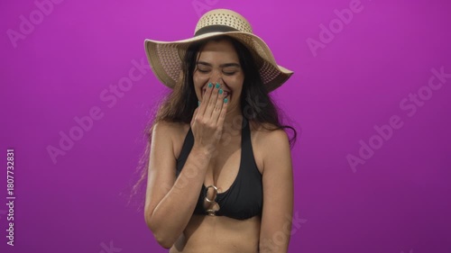 Woman wearing black bikini top and straw hat with hand covering mouth laughing in purple studio; joy.