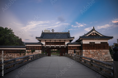 A wide wooden bridge leads to a gatehouse at Osaka Castle in Osaka, Japan at dusk. Tiled roofs and white walls stand centered as modern buildings rise beyond trees.