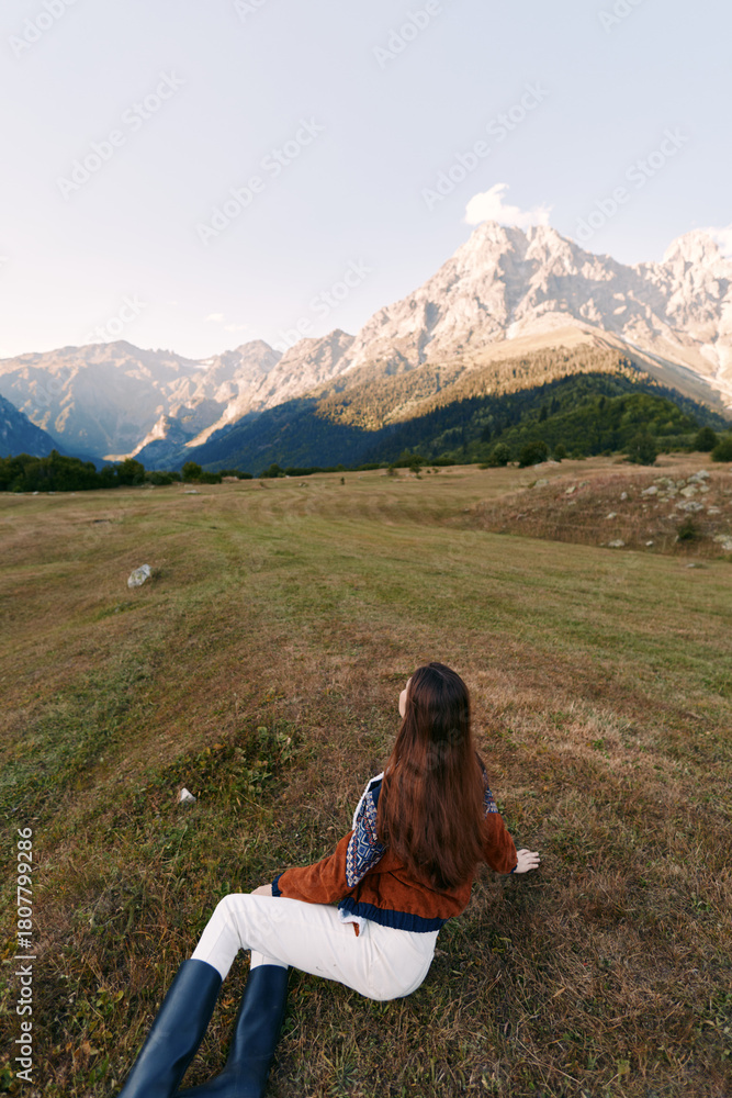 Fototapeta premium Woman lying on meadow looking at towering mountains and serene nature landscape, hiking travel scene in wide open outdoors field with boots, long hair and peaceful sky.