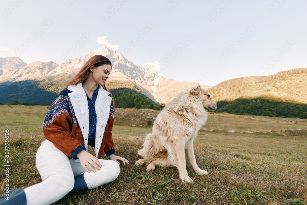 Fototapeta premium Woman and dog in mountains meadow outdoor scene, sitting and smiling as pet enjoys nature. Casual autumn clothing, relaxed pose, scenic landscape and soft natural light.
