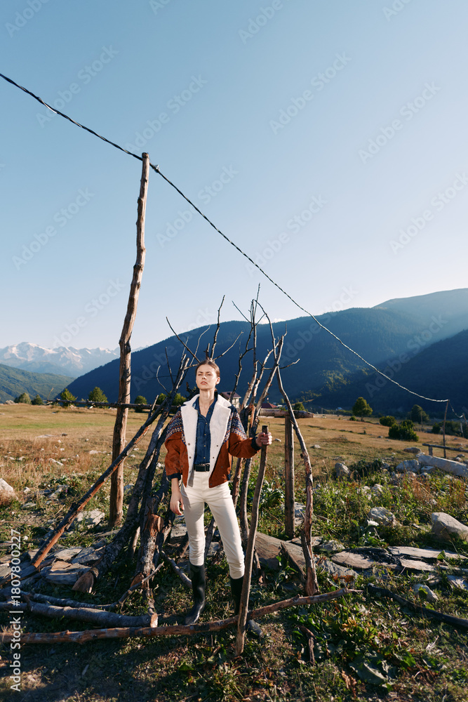 Fototapeta premium Man in traditional clothing stands by a rustic fence in a rural mountain landscape and grassy field under clear sky. Outdoor portrait, countryside lifestyle, scenic nature scene.