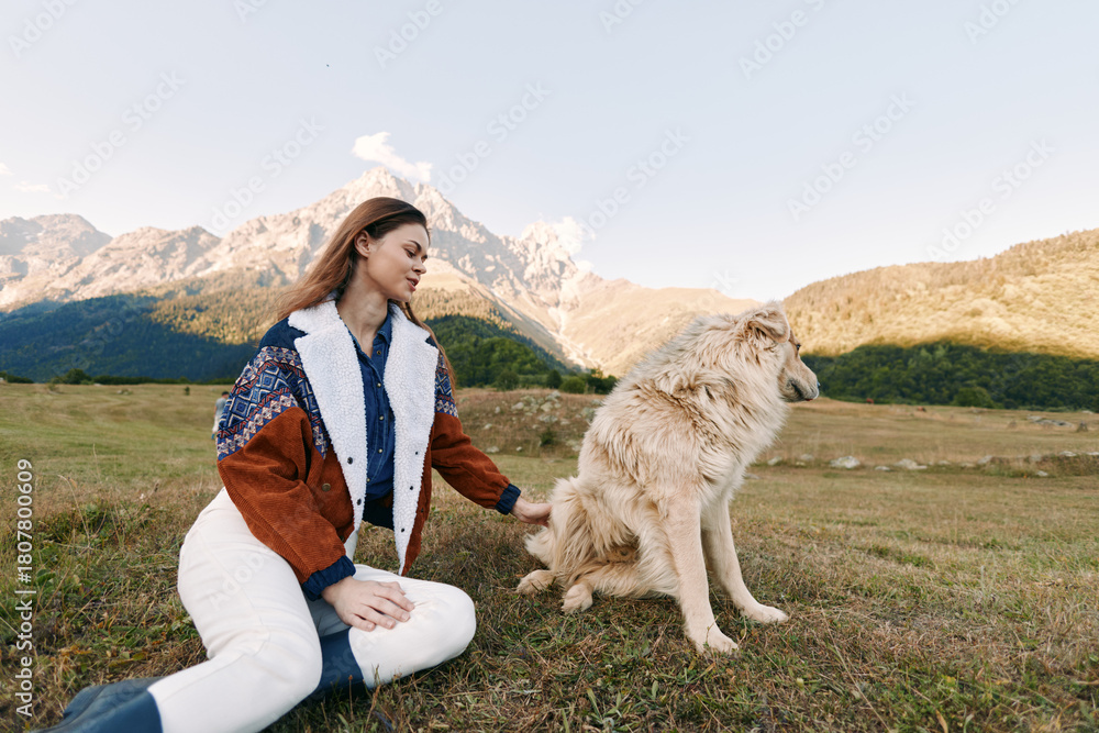 Fototapeta premium Woman and dog sitting in a mountain meadow pet portrait, outdoor nature scene with fluffy canine companion on grass, relaxed moment of bonding and scenic landscape.