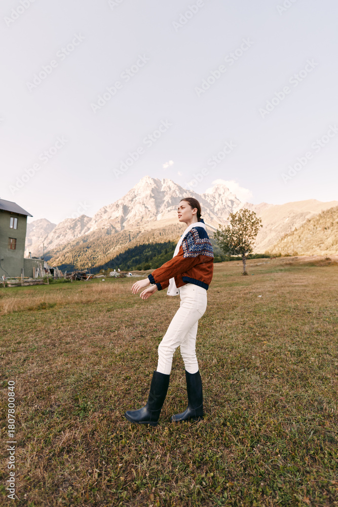 Fototapeta premium Woman walking in countryside meadow wearing a knit sweater and boots, scenic mountains in background, autumn light, rural cottage nearby, outdoor lifestyle portrait and travel vibe