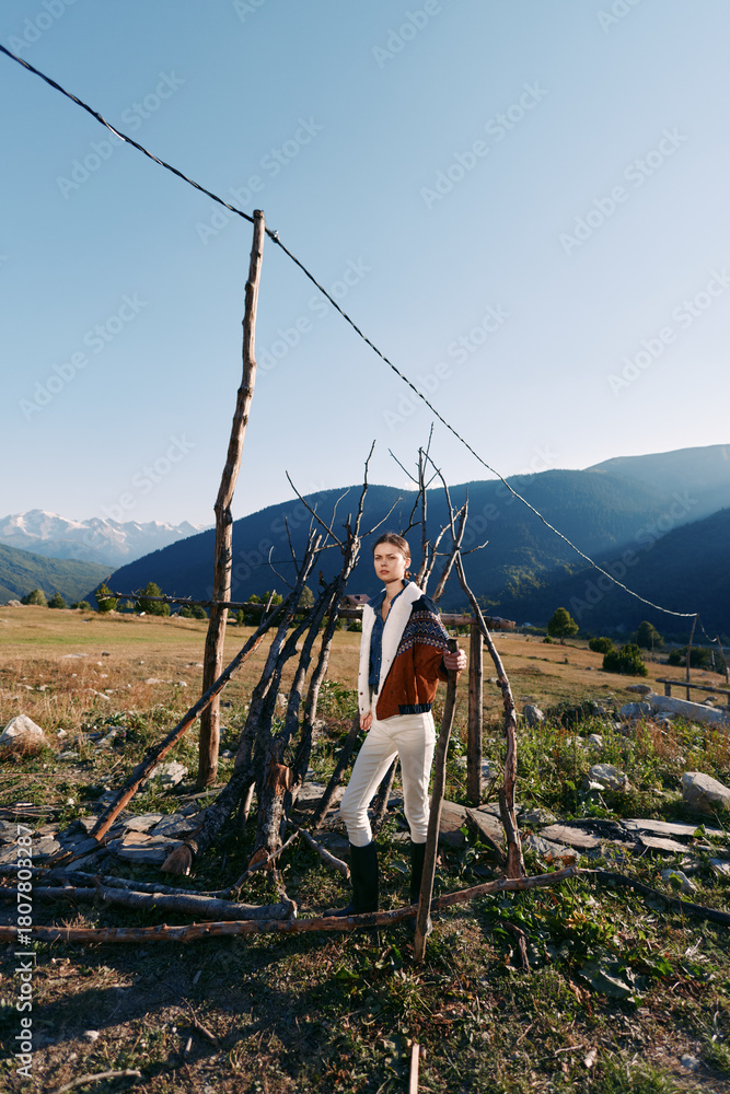 Fototapeta premium Man in traditional jacket stands by rustic fence in a rural field with mountains and wide landscape, portrait of countryside life, boots, clear sky and natural scenery.