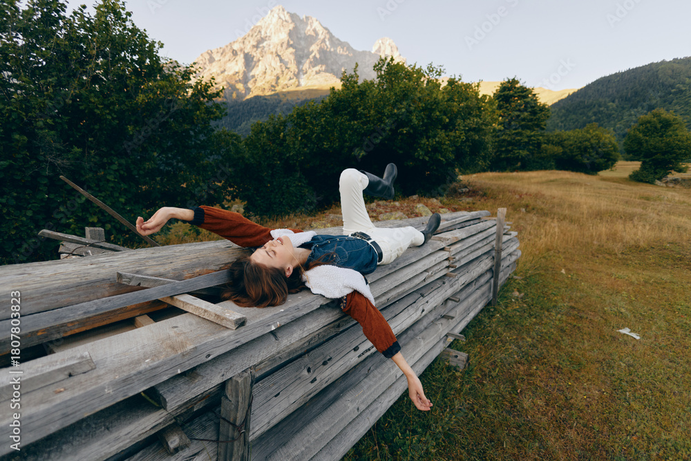 Fototapeta premium Woman lying relaxed on wooden fence in meadow with mountain backdrop, portrait of outdoors leisure and nature escape, casual clothing and serene summer landscape.