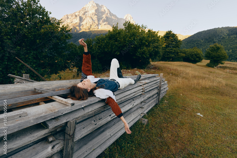 Fototapeta premium Woman on wooden fence in mountain meadow, relax outdoors among trees and nature with distant peak, casual outfit and carefree pose for peaceful escape and leisure.