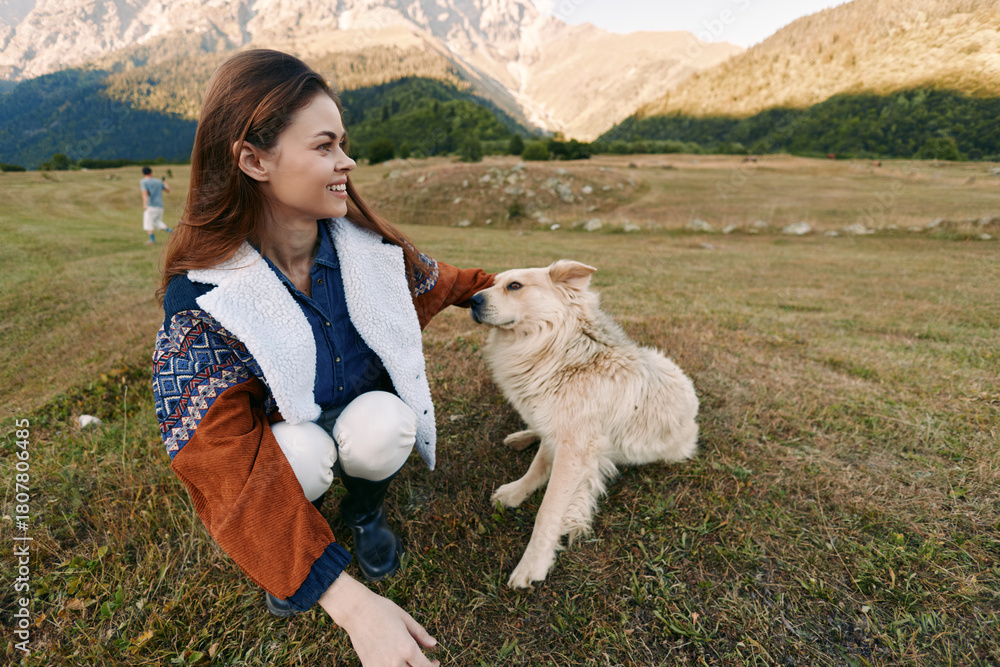 Fototapeta premium Woman crouching in a mountain field smiling while petting a dog, enjoying nature and companionship in a scenic outdoor landscape, casual jacket, happy moment with her pet