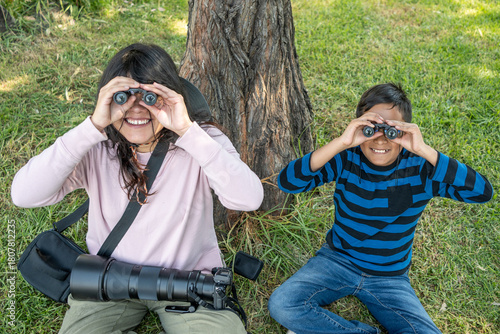 Mother and son relaxing by a tree, smiling while observing nature with binoculars