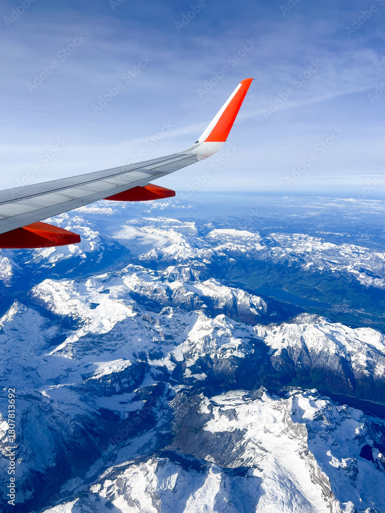Fototapeta premium Airplane wing above snow-capped mountains, showcasing breathtaking aerial view of rugged terrain, with clear blue sky and distant peaks, creating a sense of adventure and exploration