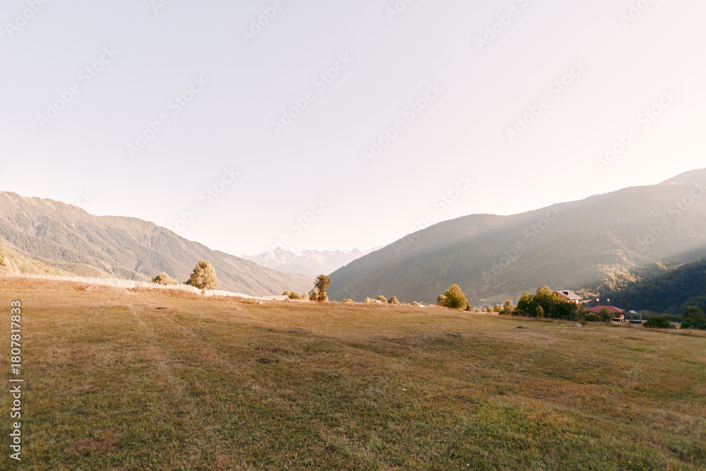 Fototapeta premium Meadow, mountains, valley, field, rural, village in a wide sunlit landscape with grassy hills and distant cottages, peaceful morning scenery for travel, hiking and countryside relaxation.