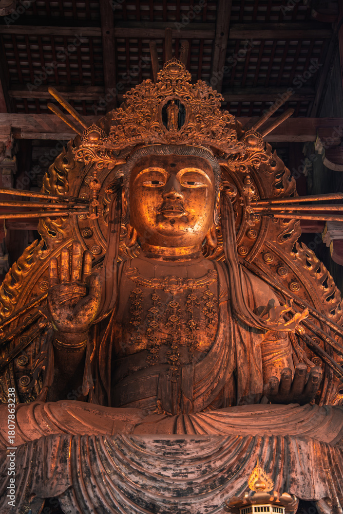 Obraz premium A large gilt Buddhist deity stands in a dim wooden hall in Nara, Japan, with a raised hand, halo like rays, crown, and layered robes lit by warm daylight.