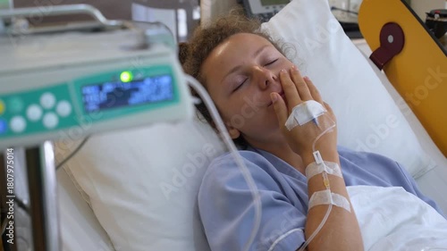 Young female patient feeling drowsy while resting in a hospital bed with an iv drip in her arm, yawning before falling asleep, showcasing the concept of medical treatment and recovery