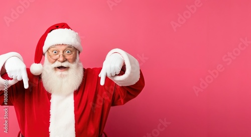 Fototapeta Naklejka Na Ścianę i Meble -  Cheerful Santa Claus in traditional red suit and hat, with a white beard and glasses, pointing downwards with both hands, standing against a bright pink studio background.