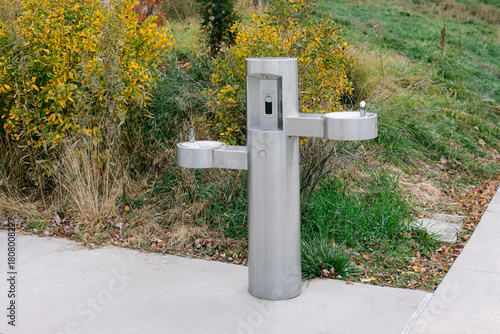 Modern stainless steel water fountain in a park surrounded by green plants and trees during a sunny day