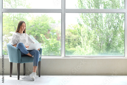 Free Download Young woman using laptop while sitting in armchair near large window in room - GFXLoad