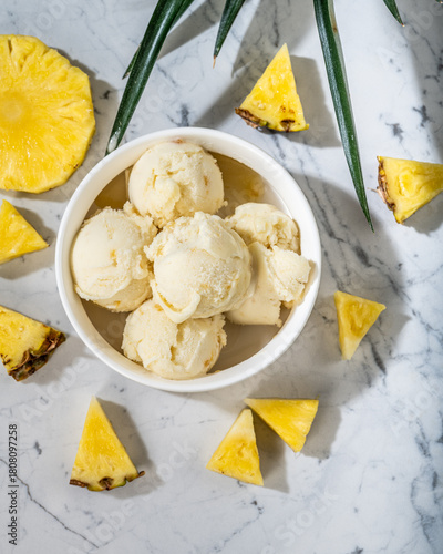 Bol de Glace à l'Ananas Frais sur Marbre. Vue aérienne d'un bol de glace crémeuse à l'ananas entouré de tranches d'ananas juteux et de feuilles sur une surface de marbre blanc.