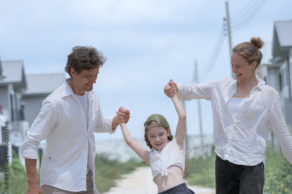 Fototapeta premium A joyful young family plays together outdoors under a bright sky. The loving parents swing their laughing daughter in the air, sharing a playful, happy moment on a summer holiday.
