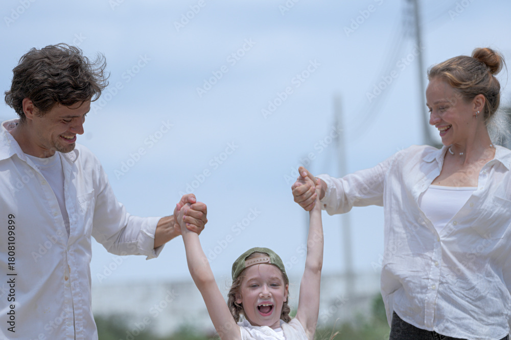 Obraz premium A joyful young family plays together outdoors under a bright sky. The loving parents swing their laughing daughter in the air, sharing a playful, happy moment on a summer holiday.