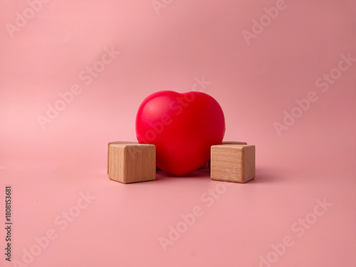 Red heart-shaped object positioned between two small wooden blocks on a pink background