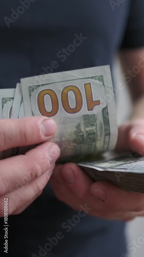 vertical screen person tallying large cash amounts casually, close view of hands and torso during bill counting activity, person in dark shirt distinctly counting hundreddollar bills