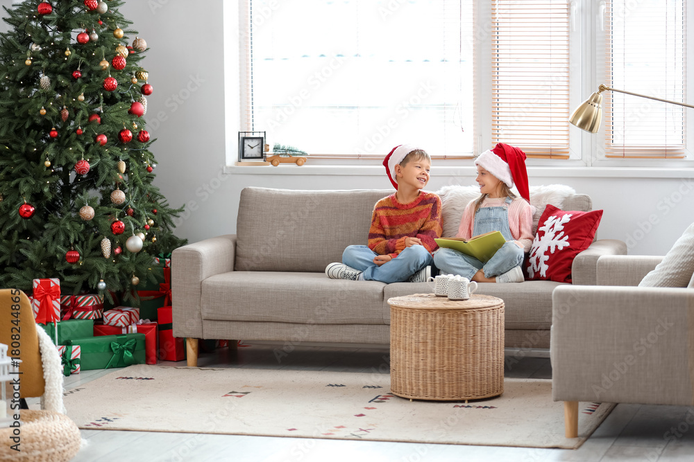 Naklejka premium Cute little children in Santa hats reading Christmas story on sofa at home