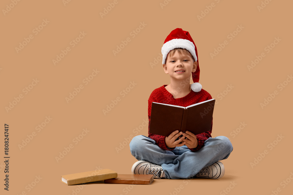 Naklejka premium Cute little boy in Santa hat reading Christmas story on beige background