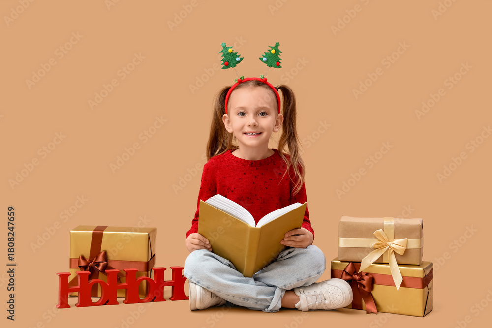 Naklejka premium Cute little girl with book and Christmas gifts sitting on beige background