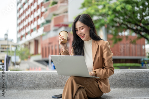 Pretty asian woman office worker holding coffee looking at laptop sitting on stone bench in a garden