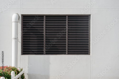 Black metal louvered ventilation grille on a white wall with red photinia leaves in the corner.