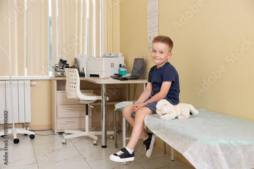 Horizontal photo. Five year old boy sits on couch during pediatrician doctor appointment, smiles, looks at camera. Children in hospital clinic office. Concept of health care, medicine, heal, work, aid