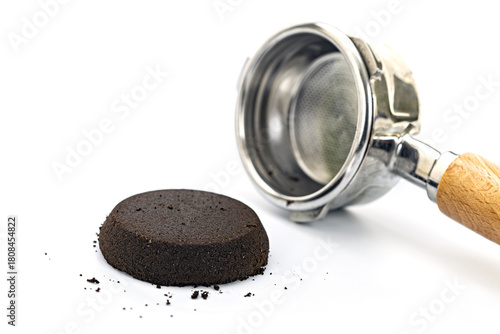 Used espresso coffee grounds puck alongside a stainless steel portafilter with wooden handle on white background