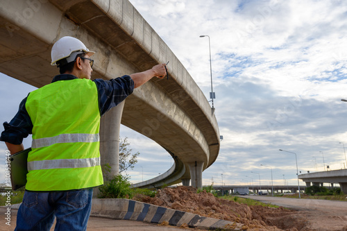 Construction engineer supervising elevated road construction, inspecting structure and directing work at infrastructure site with walkie talkie and safety gear.