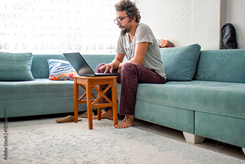 Home office working concept or sunday laziness. Man in pajamas with glasses working on laptop on coffee table. Curly haired model places online orders on computer. Comfortable, friendly atmosphere.