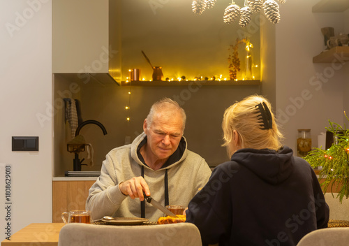 A woman and an elderly man are eating a pie, sitting at the dining table in their decorated kitchen for Christmas
