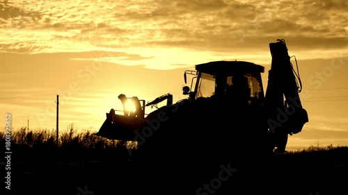 Worker working on bulldozer, backlit silhouette of powerful building machinery. Beautiful sunset or sunrise in countryside, professional dozer driver or famer, earthworks and engineering works