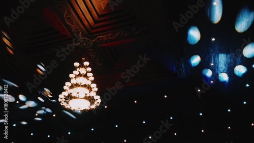 Interior of the magnificent wooden building features luxurious chandeliers and dramatic spotlights. Ceiling is decorated with carved architectural details and tiered patterns. Dynamic room atmosphere.