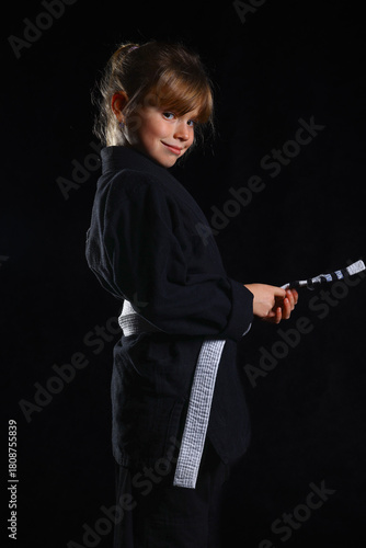 Confident young girl in black martial arts gi with white belt holding a training strap, posing proudly for kids self-defense, BJJ and martial arts lifestyle concepts.