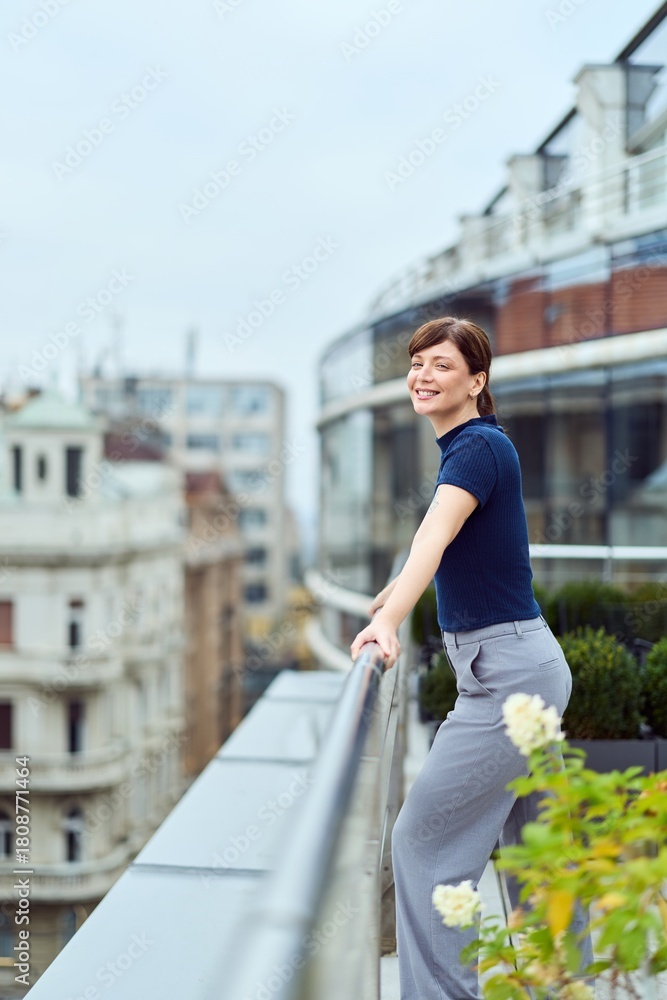 Obraz premium Smiling Woman Leaning On Balcony Rail Overlooking Cityscape In Casual Professional Outfit