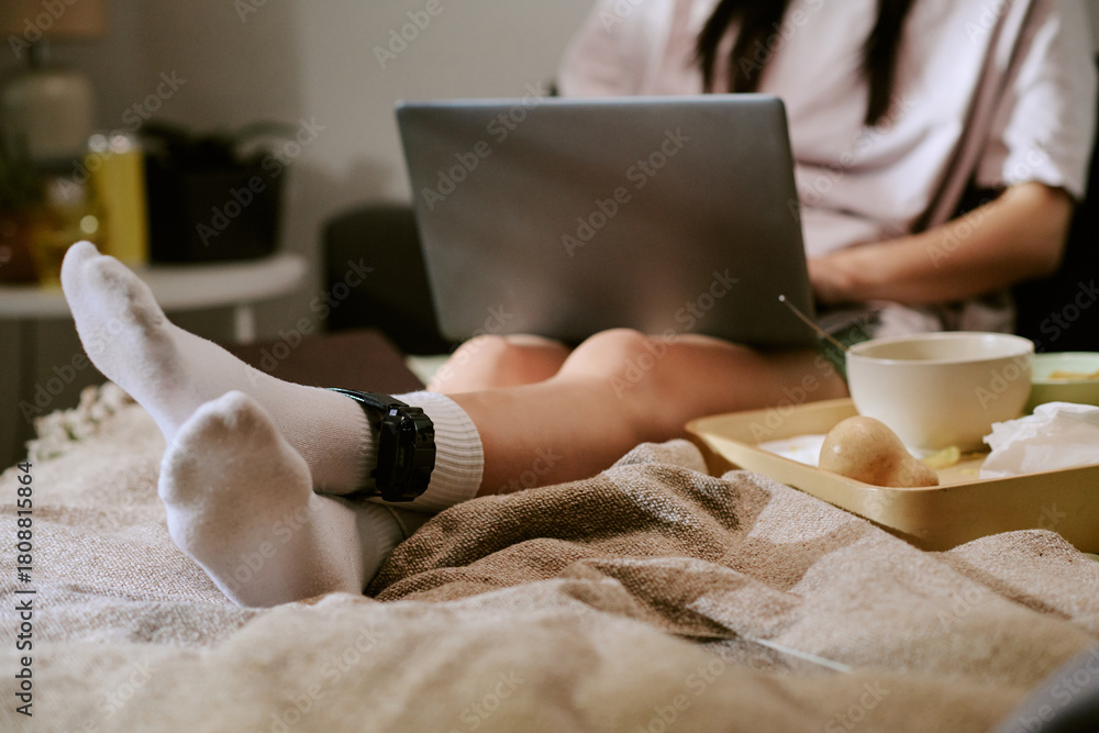 Obraz premium Unrecognizable woman sitting on bed using laptop with electronic ankle monitor visible on leg, food tray nearby, staying at home under electronic monitoring