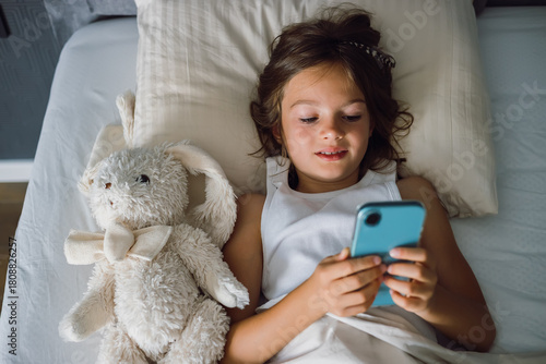 Young girl lying in bed with her plush toy, using a smartphone, illustrating modern childhood, screen time, and children's engagement with technology